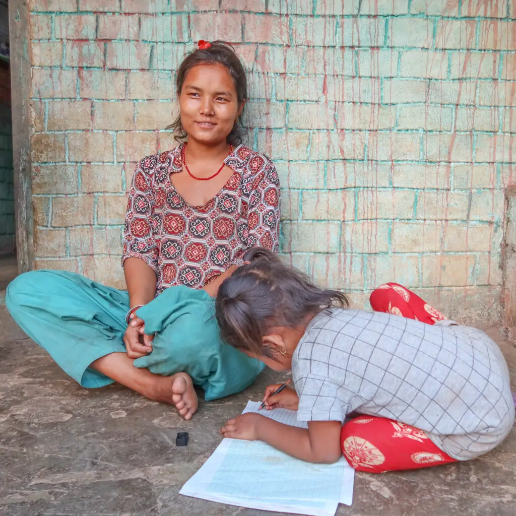 “Girl sitting outside her home in Dhading, Nepal, while a younger child writes in a notebook on the ground.