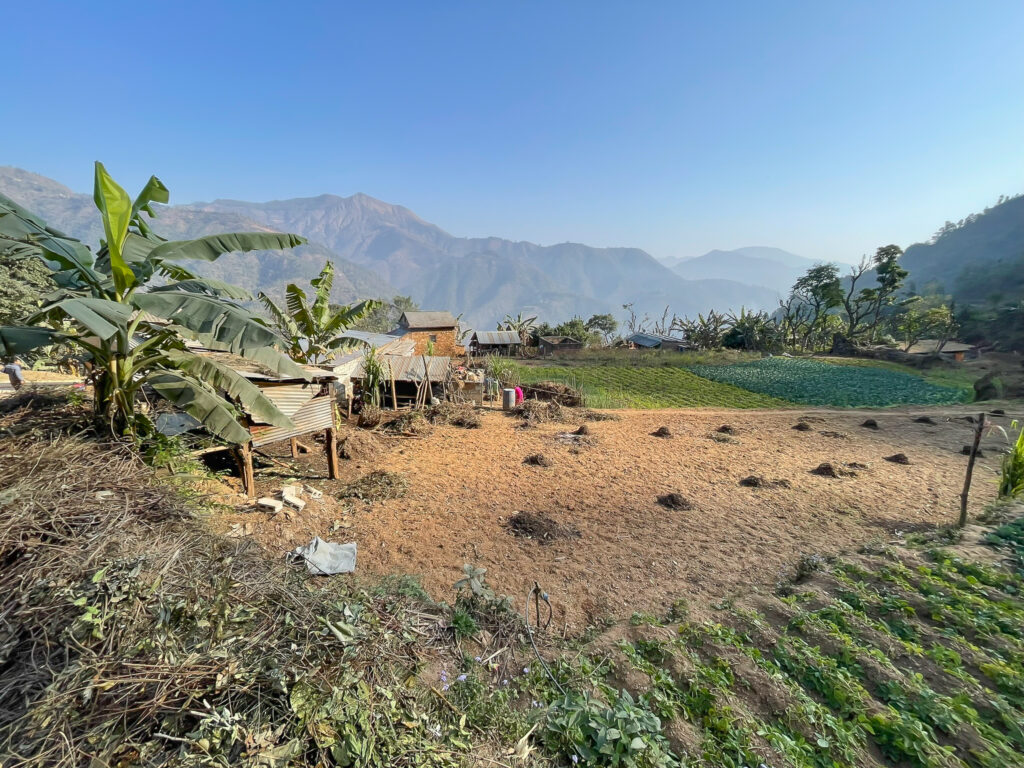 “Farmland in Dhading, Nepal, with crops, banana trees, and hillside houses against a backdrop of mountains.
