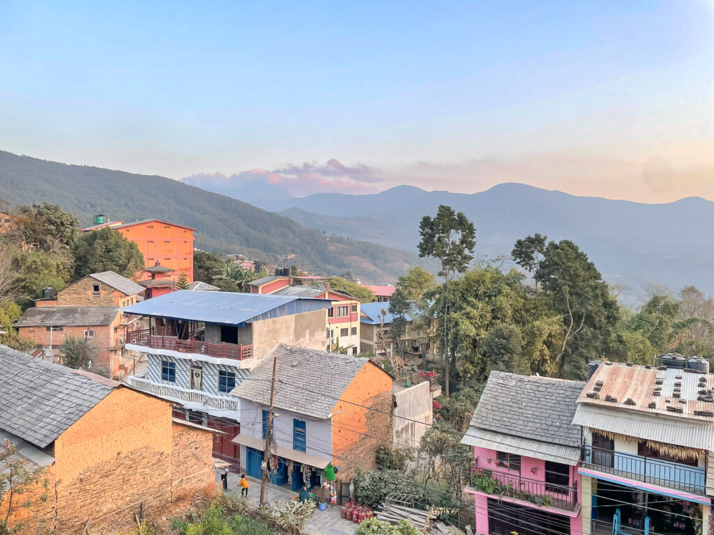 Hillside settlement in Dhading, Nepal, with brick and painted houses set against a backdrop of mountains and trees.