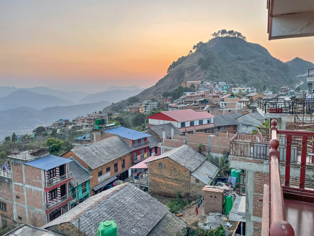 View over Dhading hillside town at sunset, with clustered houses in the foreground and mountains in the distance.