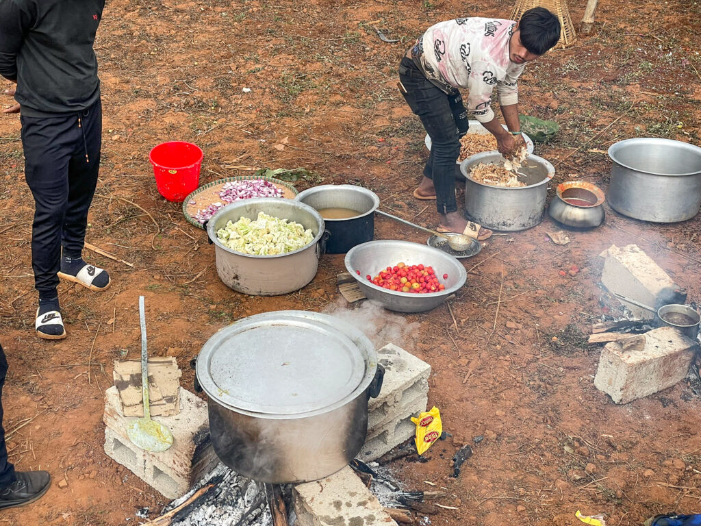 Outdoor cooking scene at the Dhading pilot site in Nepal, with large pots, chopped vegetables, and a firewood stove.