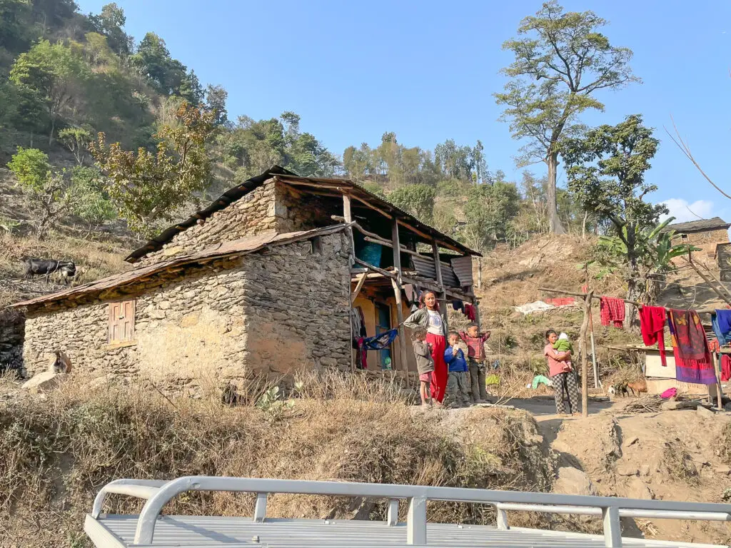 Traditional stone house in Dhading, Nepal, with family members standing outside on a sunny hillside.