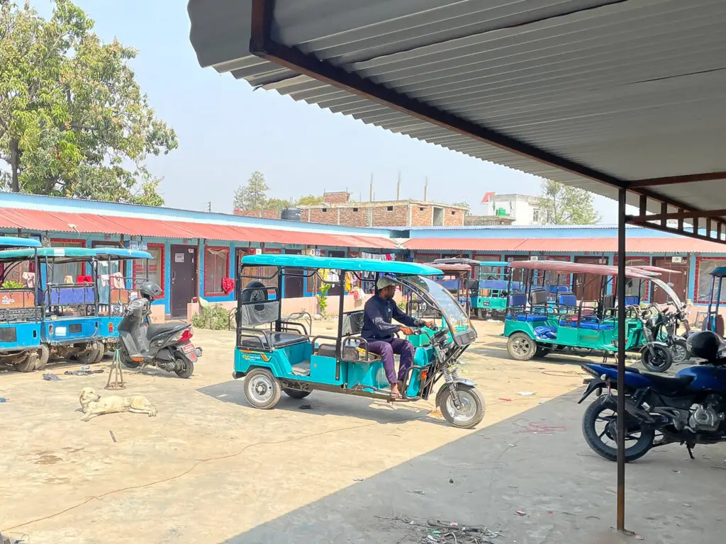 Several blue electric rickshaws parked in a courtyard in Nepal, with people nearby and motorcycles at the side.