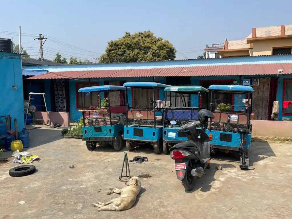 Four blue electric rickshaws parked in a courtyard in Nepal, with a scooter and dog in the foreground.