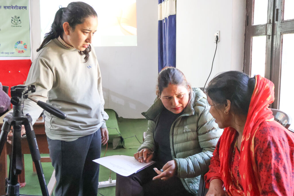 Project GANESHA team member speaking with two women indoors in Bardiya, Nepal, during a needs assessment discussion, with notes on a clipboard.”