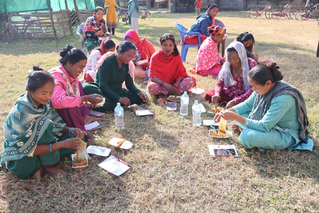 Group of women seated outdoors in Bardiya, Nepal, taking part in a small-group activity with lunch, notebooks and printed materials.