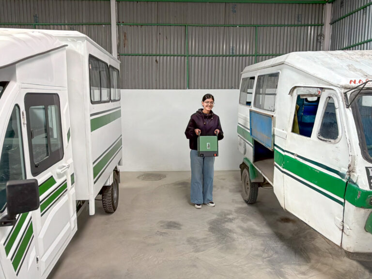 Alt 1 (best): Person holding a Project GANESHA battery module between two EV Rickshaws inside the Kathmandu facility.